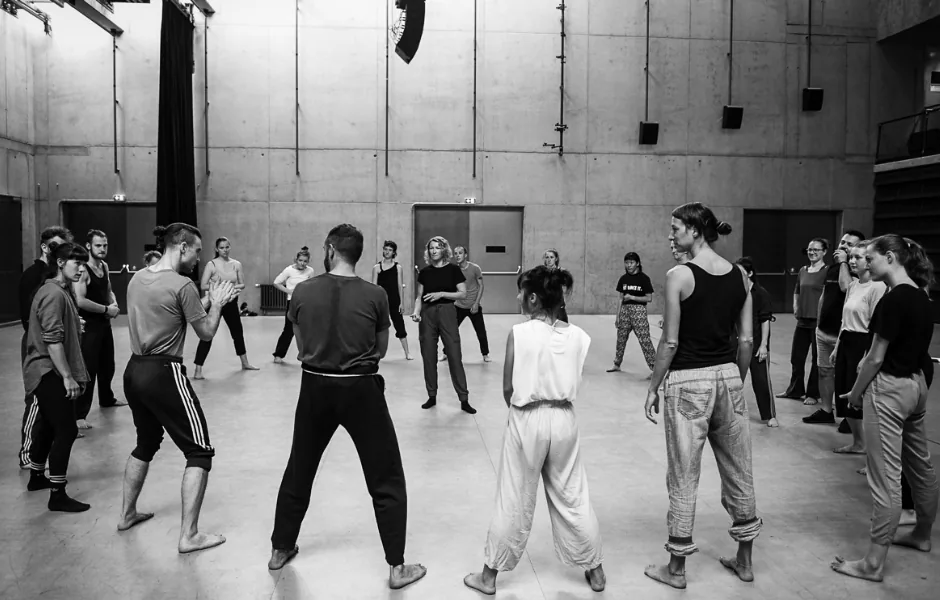 A group of people standing barefoot in a wide circle in a studio, facing each other