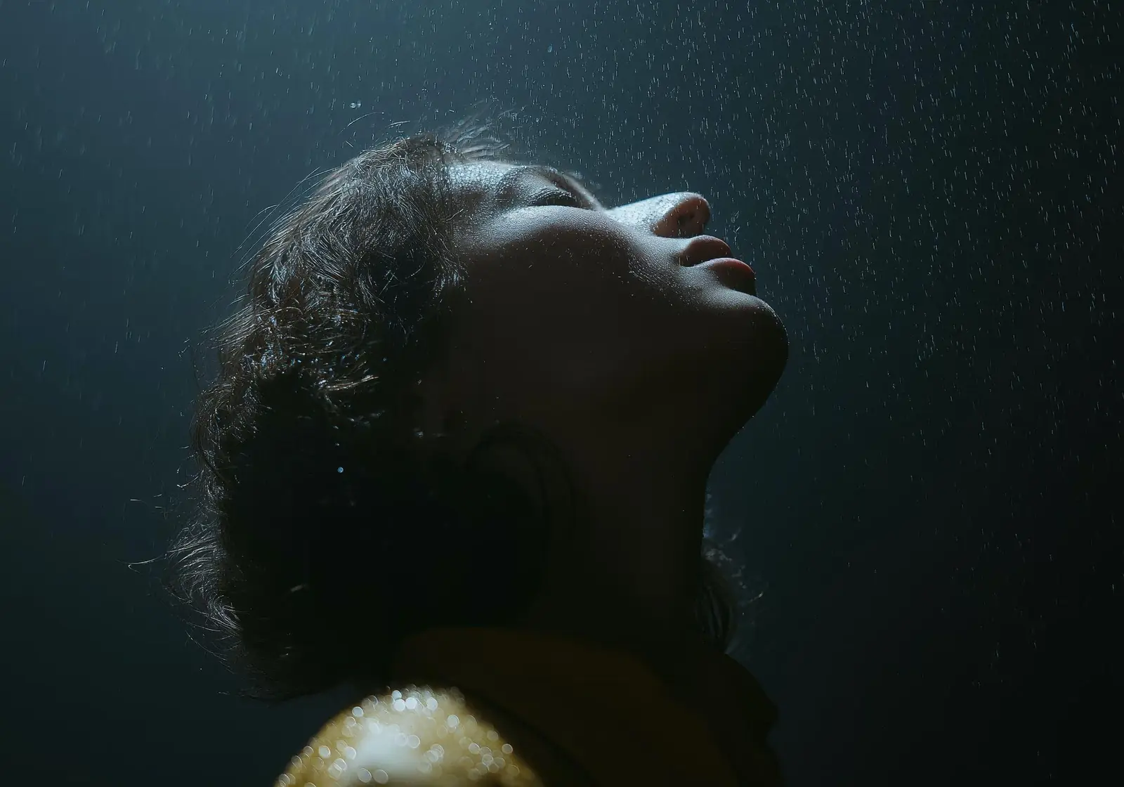 A person with eyes closed and face tilted up into falling rain, lit against a dark background