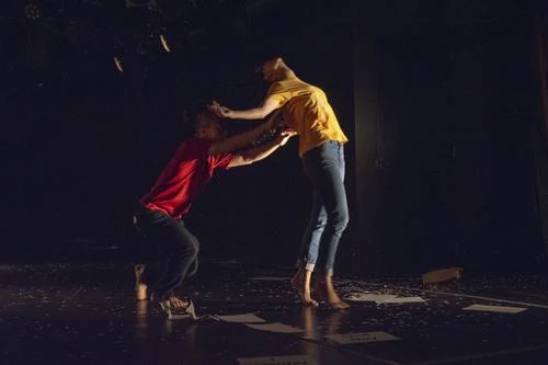 Two performers on a dark stage, one leaning back as the other reaches to hold them